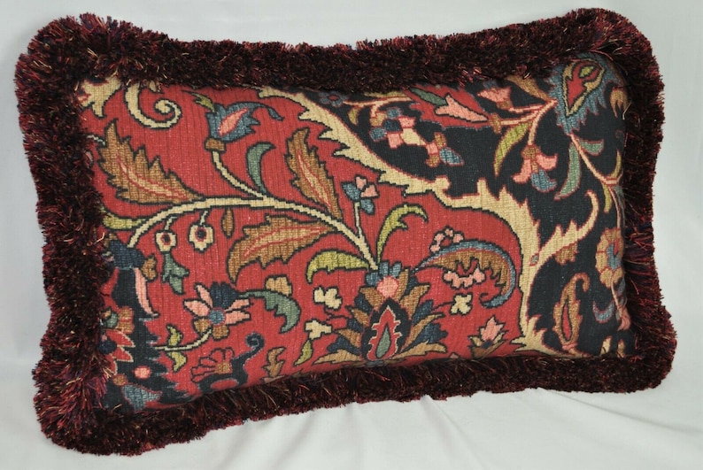 Puede incluir: Coj&iacute;n decorativo rectangular con fondo rojo y estampado floral en tonos azules, verdes y beige. El coj&iacute;n tiene un borde de flecos rojo oscuro.