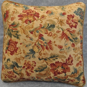 May include: A square throw pillow with a floral pattern. The pillow has a beige background with red, orange, and green flowers. The pillow has a gold trim around the edges.