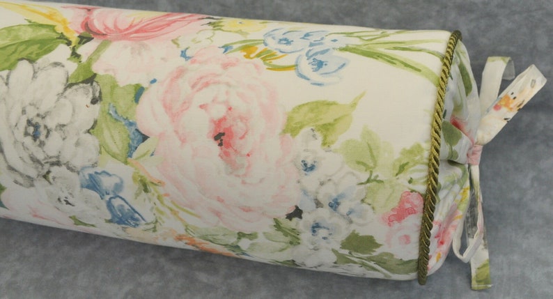 May include: A white bolster pillow with a floral print featuring pink, blue, and green watercolor flowers. The pillow has a gold trim and a white ribbon tie.