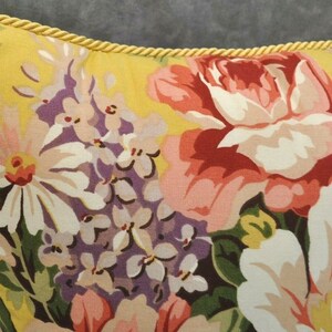 May include: A yellow pillow with a floral pattern featuring pink, red, white, and purple flowers. The pillow has a gold trim around the edges.