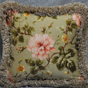 May include: A green throw pillow with a floral pattern featuring pink and yellow flowers. The pillow has a brown fringe border.