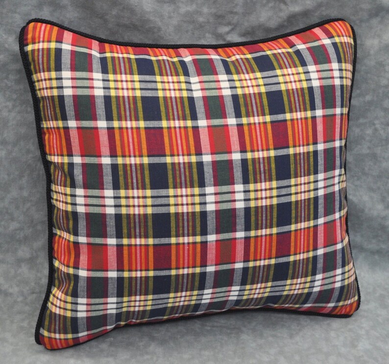 May include: A square pillow with a black trim and a plaid pattern in shades of red, yellow, blue, and green.
