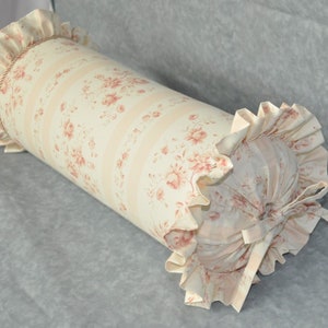 May include: A cylindrical bolster pillow with a floral pattern in pink and cream. The pillow features ruffled edges and a decorative rope detail. The fabric has vertical stripes and a repeating pattern of roses.