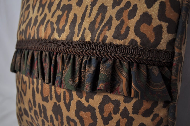 K&ouml;nnte beinhalten: Ein braunes und hellbraunes Kissen mit Leopardenmuster, braunem R&uuml;schenbesatz und darunterliegendem Stoff mit Paisley-Muster.