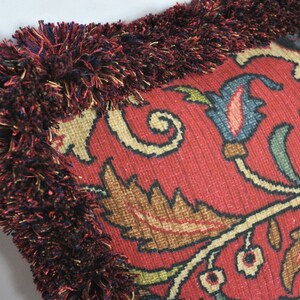Puede incluir: Primer plano de un coj&iacute;n decorativo con fondo rojo y un intrincado dise&ntilde;o floral en tonos beige, marr&oacute;n y azul. El coj&iacute;n est&aacute; adornado con un grueso borde con flecos en rojo oscuro e hilos multicolores.