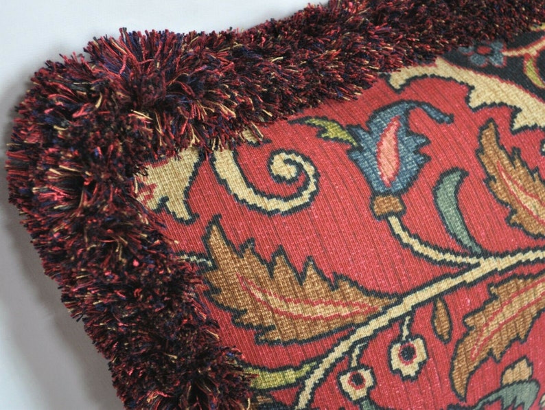 Puede incluir: Primer plano de un coj&iacute;n decorativo con fondo rojo y un intrincado dise&ntilde;o floral en tonos beige, marr&oacute;n y azul. El coj&iacute;n est&aacute; adornado con un grueso borde con flecos en rojo oscuro e hilos multicolores.