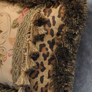May include: A decorative pillow with a brown and tan leopard print fabric, a floral patterned fabric, and gold tassels. The pillow has a brown fringe border.