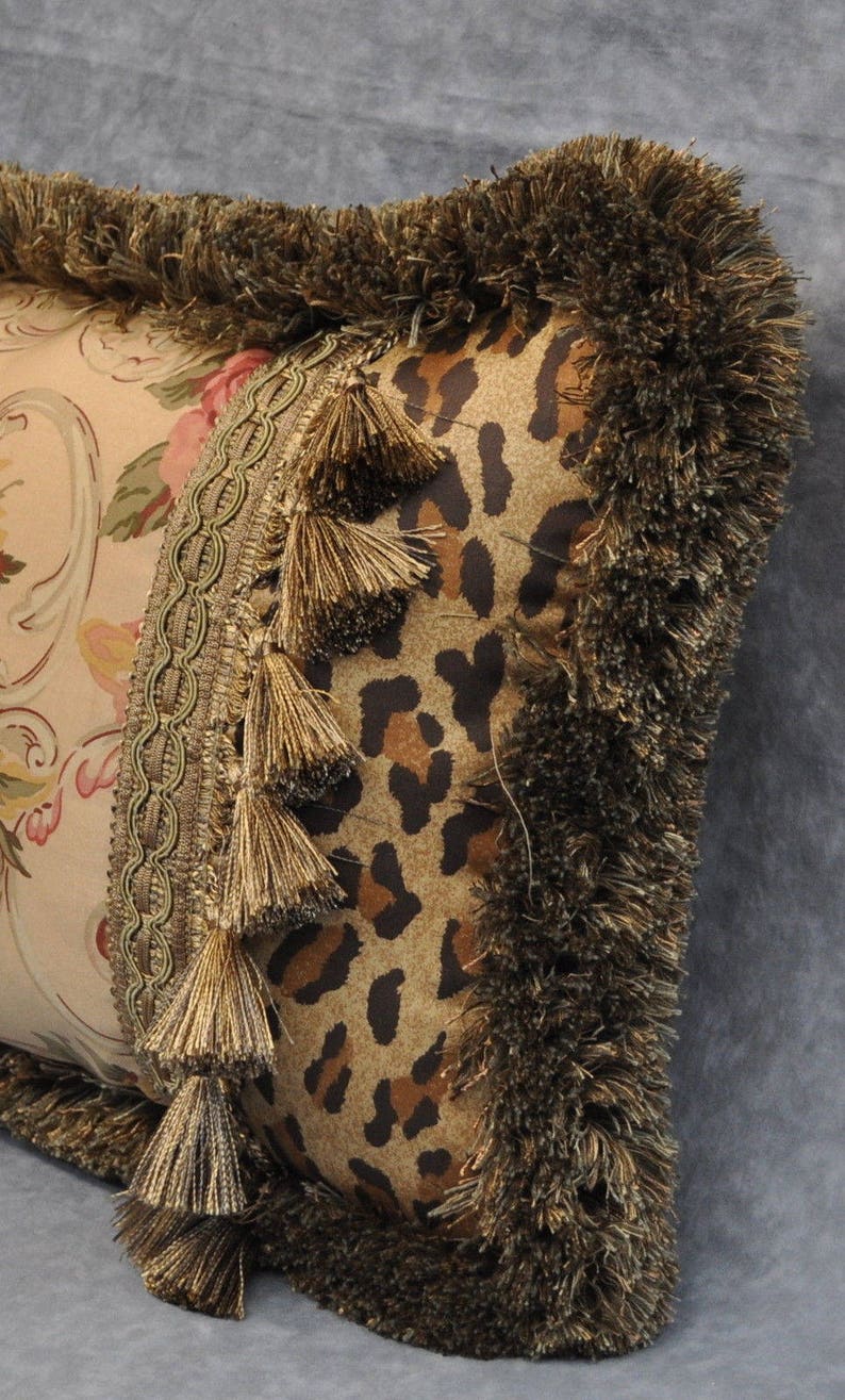May include: A decorative pillow with a brown and tan leopard print fabric, a floral patterned fabric, and gold tassels. The pillow has a brown fringe border.