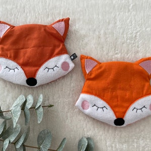 Könnte beinhalten: Zwei orangefarbene Kissen in Form eines Fuchses mit weißen Gesichtern und rosa Ohren. Die Kissen haben gestickte Augen, eine schwarze Nase und einen weißen Mund.