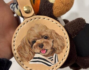 May include: A round leather keychain with a painted portrait of a brown poodle wearing a striped shirt. The keychain has a gold metal ring and a brown leather strap.