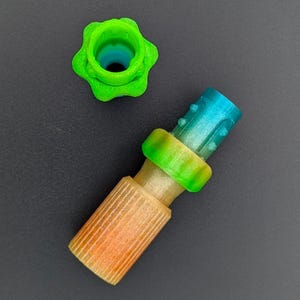 May include: A disassembled, multi-coloured 3D-printed object, possibly a connector or fitting. The components include a green, star-shaped piece, and a cylindrical piece with orange, green, and blue sections. The parts are arranged against a dark grey background.