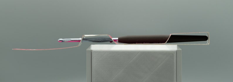 Peut inclure: Un crochet en argent avec une poign&eacute;e noire est pr&eacute;sent&eacute; sur un support blanc. Le crochet est plac&eacute; dans un support en plastique transparent.