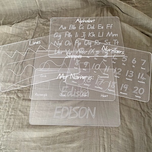 May include: Four clear plastic dry-erase boards with white lines and text. The boards include an alphabet, numbers 1-20, shapes, and lines. The text "My Name is: Edison" is written on the shapes board.