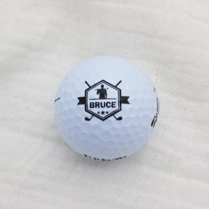 May include: White golf ball with a black logo featuring a golfer silhouette, the name "BRUCE" and three stars.