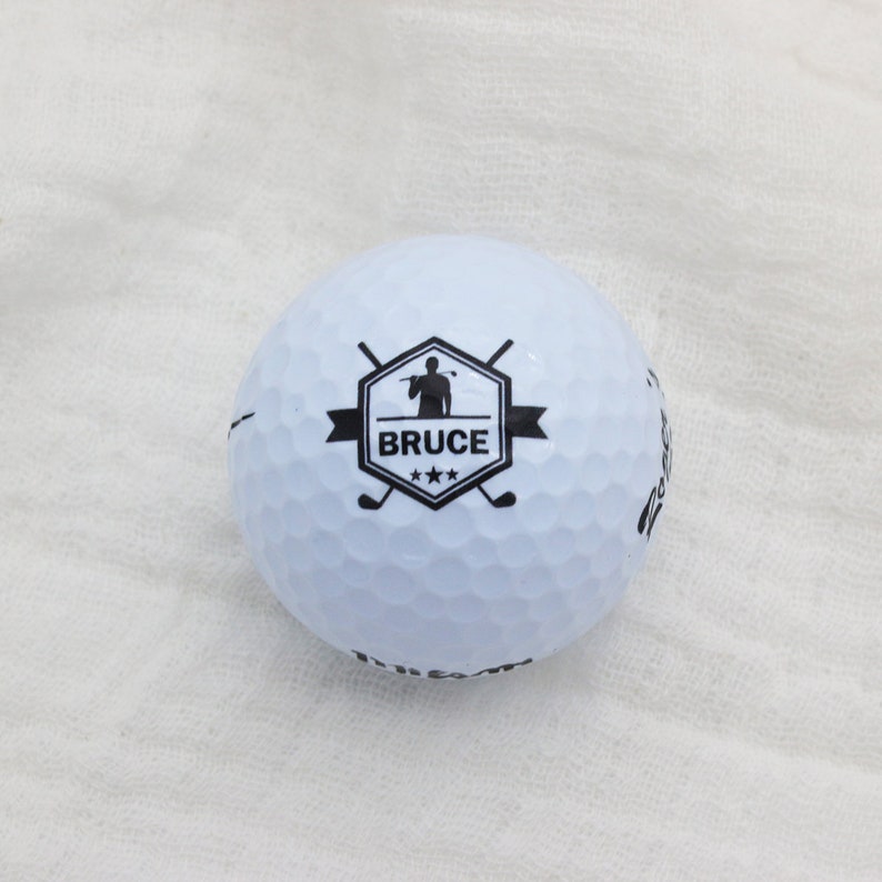 May include: White golf ball with a black logo featuring a golfer silhouette, the name "BRUCE" and three stars.