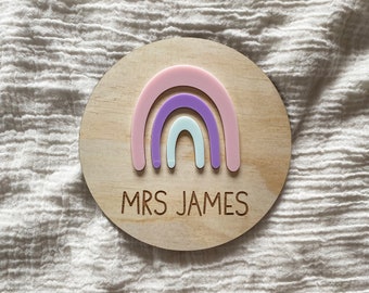 May include: A circular wooden plaque featuring a pastel rainbow in pink, lilac, and pale blue. The name "MRS JAMES" is engraved beneath the rainbow. The plaque is set against a white, textured backdrop.