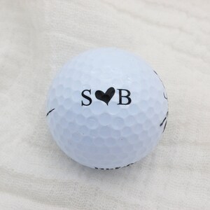 May include: A white golf ball with black lettering that says "S&hearts;B" on it.