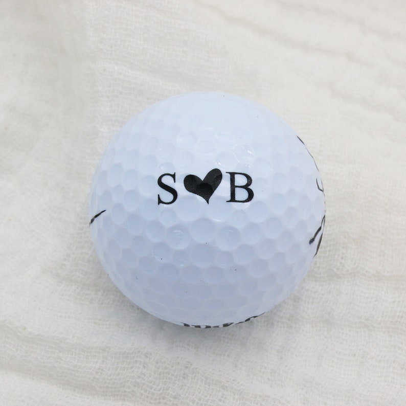 May include: A white golf ball with black lettering that says "S&hearts;B" on it.