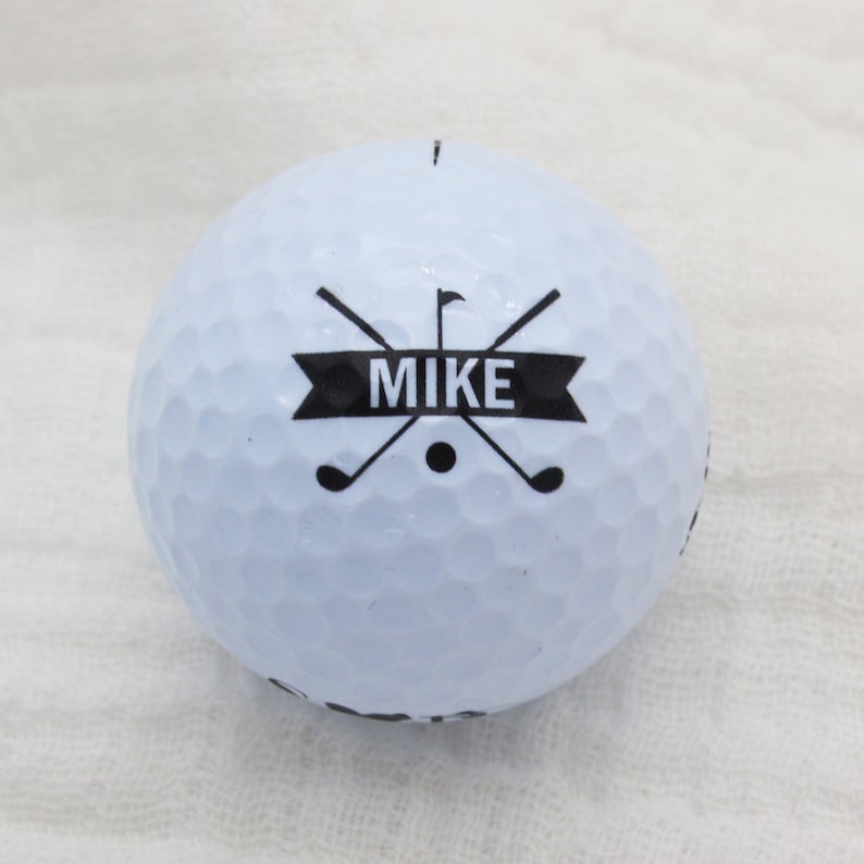 May include: White golf ball with a black design featuring crossed golf clubs and a banner with the name "MIKE" printed on it.