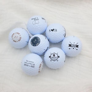 May include: Seven white golf balls with black printed designs. The designs include names, initials, and a golf club logo. The text on the balls reads "Roger's Balls", "SB", "Mike", "Bruce", "ITCH + IMK", and "ITCH + IMK".