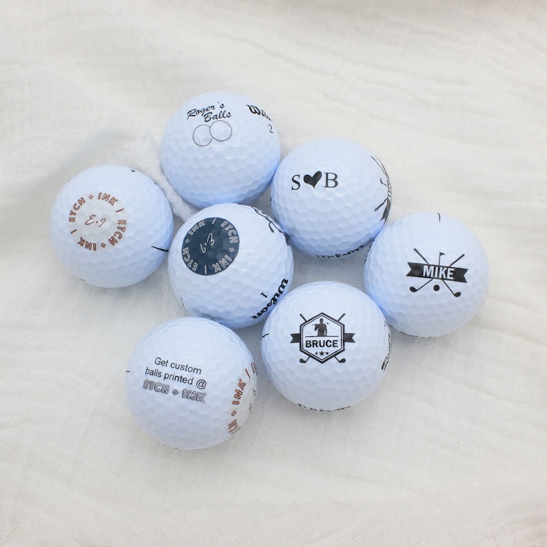 May include: Seven white golf balls with black printed designs. The designs include names, initials, and a golf club logo. The text on the balls reads "Roger's Balls", "SB", "Mike", "Bruce", "ITCH + IMK", and "ITCH + IMK".