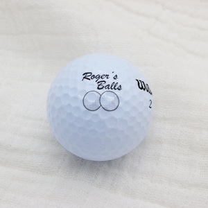 May include: A white golf ball with the text "Roger's Balls" and two circles printed on it. The ball is a Wilson brand.