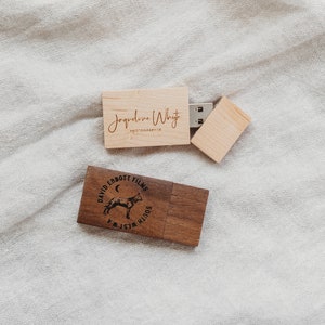 Op de afbeelding: Twee houten USB-sticks. Eén is lichtbruin met de tekst "Jacqueline Whyte Photographer" erop gegraveerd. De andere is donkerbruin met een cirkelvormig ontwerp met een wolf die huilt naar de maan, de tekst "David Ebbott Films" en de woorden "South West WA".
