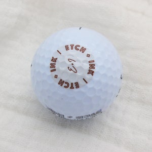 May include: A white golf ball with a brown circle on it that says "ETCH x INK" and "ETCH x INK" with a handwritten "3" in the center.