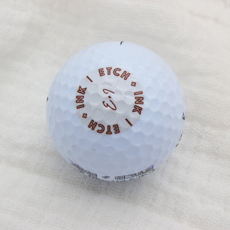 May include: A white golf ball with a brown circle on it that says "ETCH x INK" and "ETCH x INK" with a handwritten "3" in the center.