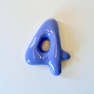 May include: A glossy, periwinkle blue ceramic number four with a smooth, rounded, and inflated appearance. The digit features a central open loop and a reflective surface that catches light, creating bright white highlights across its curved form.