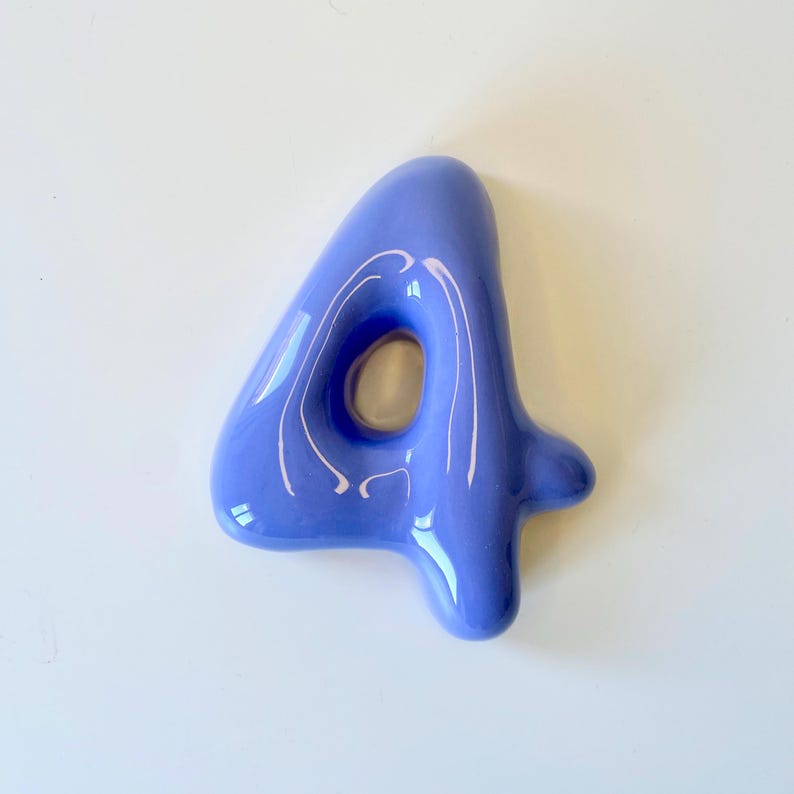 May include: A glossy, periwinkle blue ceramic number four with a smooth, rounded, and inflated appearance. The digit features a central open loop and a reflective surface that catches light, creating bright white highlights across its curved form.