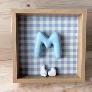 Può includere: Una cornice quadrata in legno chiaro contenente una lettera M azzurra centrata sopra una piccola statuetta di orso polare bianco e un oggetto bianco a forma di cuore, il tutto su uno sfondo con motivo a quadretti azzurro e bianco.