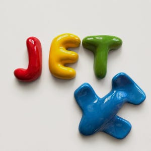 May include: A set of glossy, rounded ceramic or plastic letters spelling the word JET in red, yellow, and green, accompanied by a separate blue airplane-shaped figure. Each piece has a smooth, reflective surface and a three-dimensional shape.