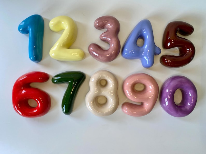 May include: A set of ten glossy, rounded ceramic numbers from zero to nine, each featuring a distinct solid colour including blue, yellow, mauve, periwinkle, dark brown, red, forest green, cream, light pink, and purple, arranged in two rows.