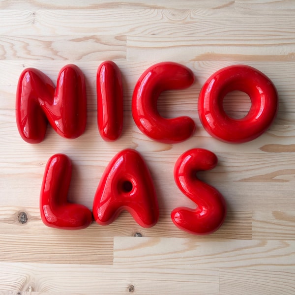 May include: A set of glossy, bright red, three-dimensional balloon-style letters spelling out the name NICOLAS. Each letter has a smooth, rounded, inflated appearance with a reflective surface, arranged in two rows on a light wood grain surface.
