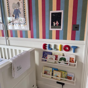 K&ouml;nnte beinhalten: Ein wei&szlig;es Kinderbett mit einer wei&szlig;en Decke, die mit dem Namen Eli bestickt ist, neben einer Wand mit vertikal gestreifter Tapete, zwei wei&szlig;en schwebenden B&uuml;cherregalen mit Kinderb&uuml;chern und dem Namen ELLIOT in bunten, abgerundeten Buchstaben.