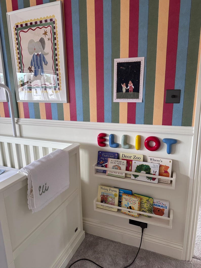 K&ouml;nnte beinhalten: Ein wei&szlig;es Kinderbett mit einer wei&szlig;en Decke, die mit dem Namen Eli bestickt ist, neben einer Wand mit vertikal gestreifter Tapete, zwei wei&szlig;en schwebenden B&uuml;cherregalen mit Kinderb&uuml;chern und dem Namen ELLIOT in bunten, abgerundeten Buchstaben.