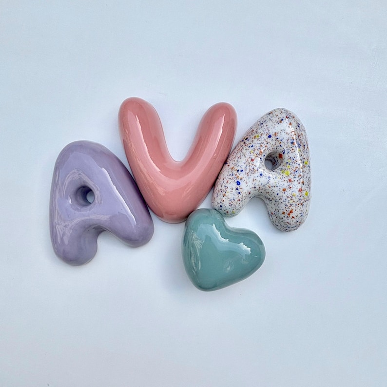 May include: Four ceramic decorative pieces spelling the name "AVA". The letters are in a lavender, pink, and speckled white finish. A small, glossy, teal heart is also included in the arrangement.