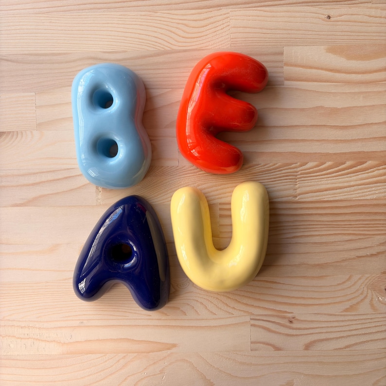 May include: Four individual ceramic letters arranged in a square formation. The top left is a light blue letter B, the top right is a bright orange letter E, the bottom left is a dark navy blue letter A, and the bottom right is a pale yellow letter U.
