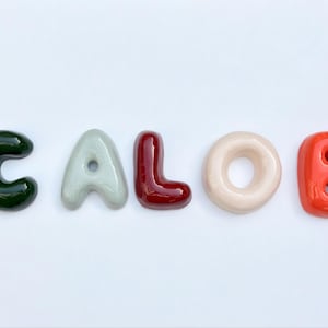 May include: A collection of glossy, colourful letters spelling "CALOB" against a white backdrop. The letters are in shades of green, grey, burgundy, cream, and orange-red, creating a vibrant and appealing visual.