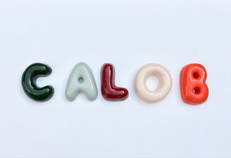 May include: A collection of glossy, colourful letters spelling "CALOB" against a white backdrop. The letters are in shades of green, grey, burgundy, cream, and orange-red, creating a vibrant and appealing visual.