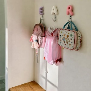 May include: Three wall-mounted metal hooks topped with letter-shaped decor spelling EAR in lavender, speckled white, and pink. Hanging from the hooks are a metallic pink backpack, a pink dress with a white collar, and a rectangular patterned bag.