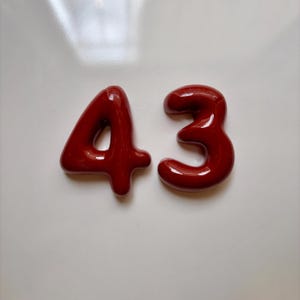 May include: Two glossy, dark red, three-dimensional numbers representing the number 43. Each digit features a smooth, rounded, bulbous shape with a reflective surface finish, creating a bold and distinct appearance against a plain white background.