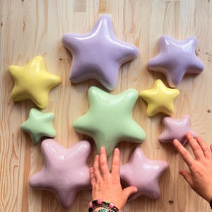 Op de afbeelding: Een verzameling van negen gladde, glanzende, stervormige keramische objecten in diverse pastelkleuren, waaronder lichtpaars, lichtgeel, mintgroen en zachtroze, gerangschikt in een groep op een lichtgekleurd houten oppervlak met zichtbare nerf.