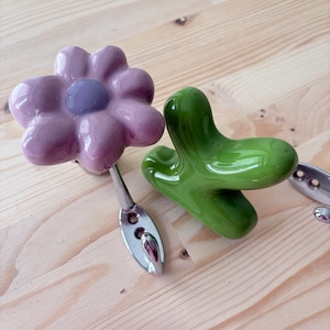 Op de afbeelding: Twee wandhaken met decoratieve keramische knoppen. De ene haak heeft een glanzende lavendelkleurige bloemvorm met een donkerpaars centrum. De andere haak heeft een gladde, glanzende, felgroene abstracte vorm. Beide haken hebben zilverkleurige metalen voetjes.