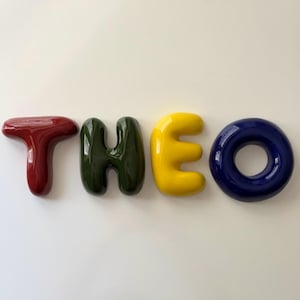 K&ouml;nnte beinhalten: Vier einzelne, gl&auml;nzende, dreidimensionale Buchstaben, die den Namen THEO bilden. Der Buchstabe T ist dunkelrot, das H ist dunkelgr&uuml;n, das E ist hellgelb und das O ist tief dunkelblau. Jeder Buchstabe hat eine glatte, abgerundete, aufgebl&auml;hte Optik.