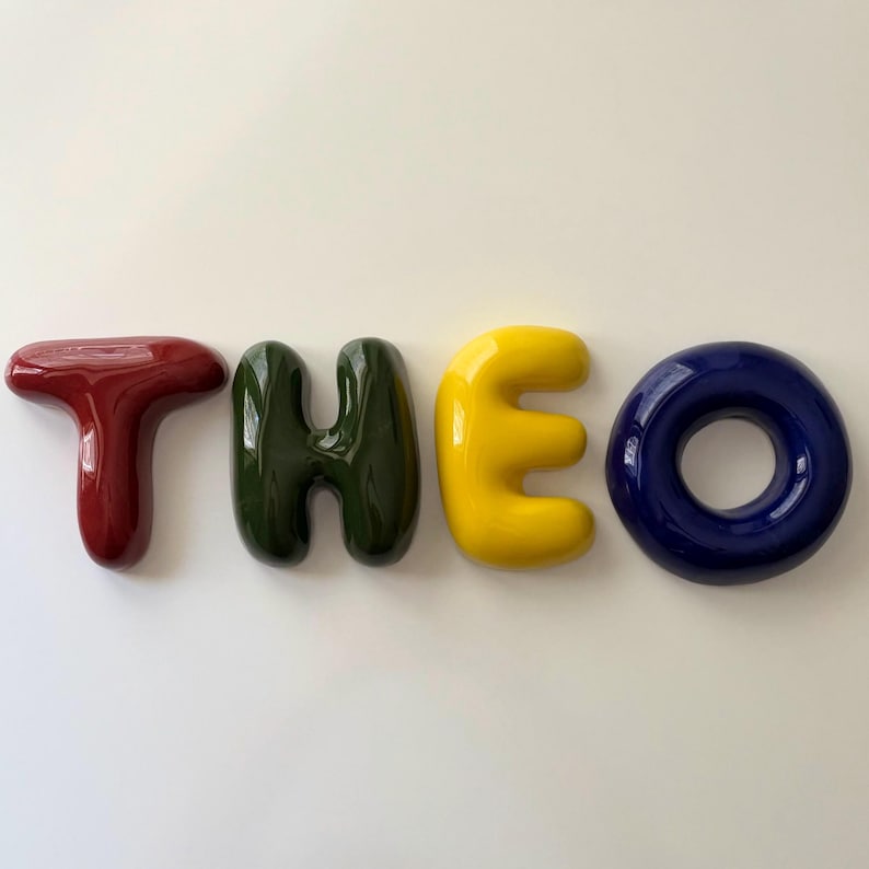 K&ouml;nnte beinhalten: Vier einzelne, gl&auml;nzende, dreidimensionale Buchstaben, die den Namen THEO bilden. Der Buchstabe T ist dunkelrot, das H ist dunkelgr&uuml;n, das E ist hellgelb und das O ist tief dunkelblau. Jeder Buchstabe hat eine glatte, abgerundete, aufgebl&auml;hte Optik.