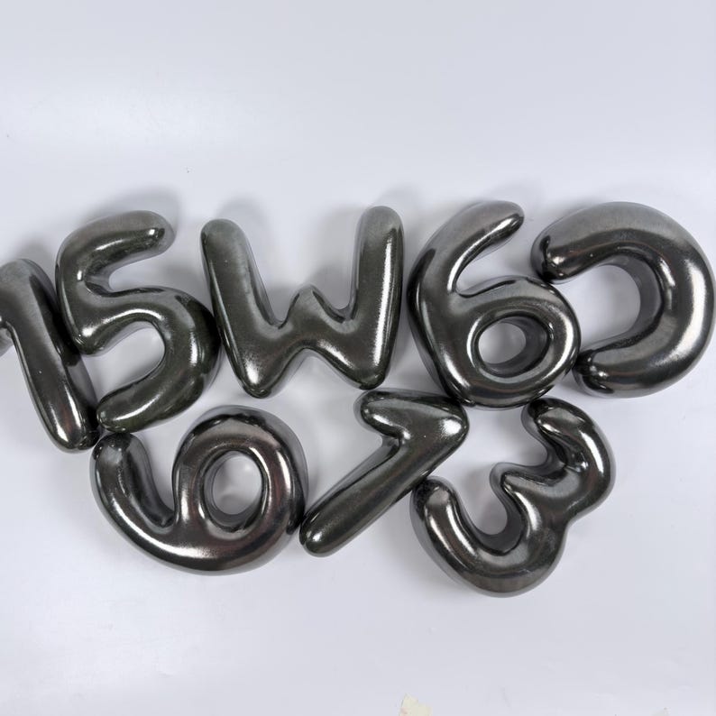 Może przedstawiać: Zestaw siedmiu pojedynczych metalicznych srebrnych cyfr i liter w stylu balonowym, w tym cyfry 1, 5, 6, 3, 9, 7 oraz litera W, wszystkie o gładkim, odblaskowym i zaokrąglonym wyglądzie przypominającym nadmuchane kształty.