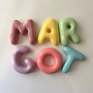 May include: A set of six individual, glossy, balloon-style letters spelling the name MARGOT. The letters are arranged in two rows, featuring a mauve M, yellow A, light green R, lavender G, coral O, and light blue T, all with a smooth finish.