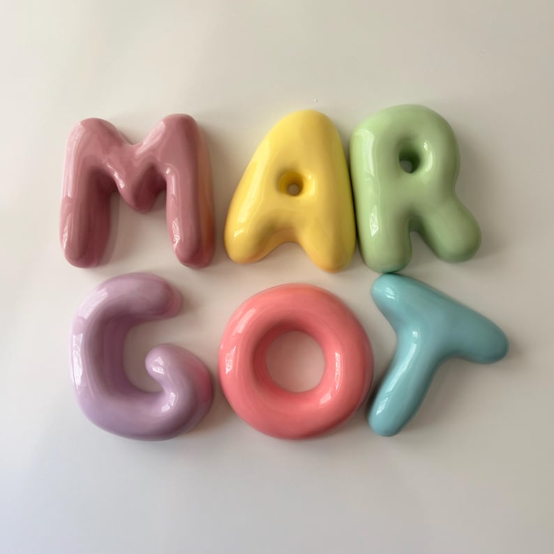 May include: A set of six individual, glossy, balloon-style letters spelling the name MARGOT. The letters are arranged in two rows, featuring a mauve M, yellow A, light green R, lavender G, coral O, and light blue T, all with a smooth finish.